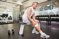 A man with a knee Injury at the gym sitting on a workout bench holding his knee which is wrapped in bandages. 